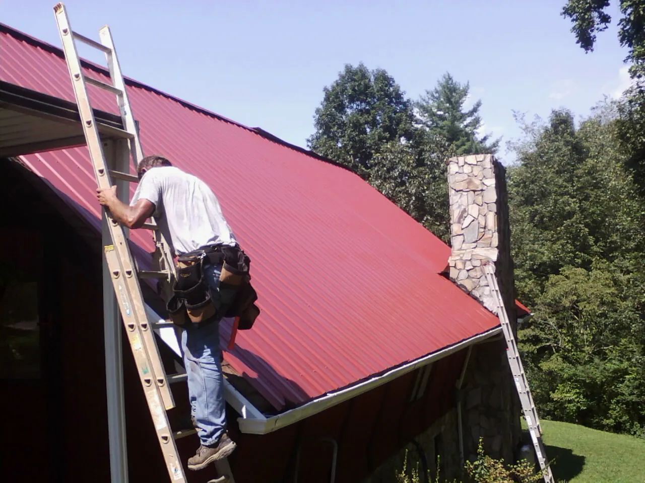 Roofer on a ladder beside a red metal roof and stone chimney — Soffit & Fascia Repair by Roofing Brothers in Niceville